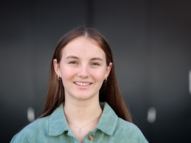 Portrait einer lachenden, jungen Frau, die ein grünes Overshirt trägt und vor einem schwarzen Hintergrund steht. 