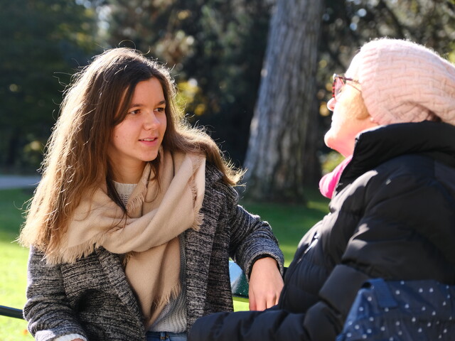 Eine junge Frau, die einen beigen Schal und grauen Wintermantel trägt, sitzt einer Seniorin mit schwarzer Winterjacke und rosa Strickmütze auf einer Parkbank gegenüber.