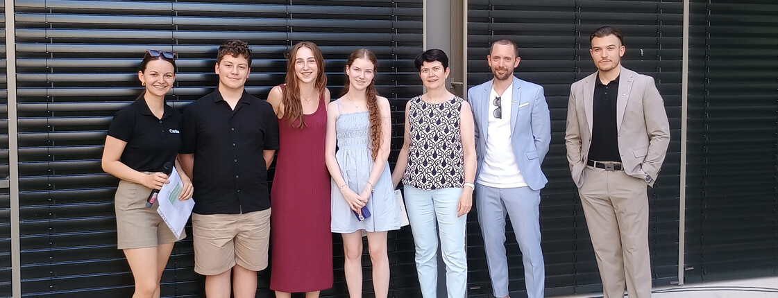 Sieben Personen stehen nebeneinander vor den schwarzen Schalusien einer Hausfassade zu einem Gruppenfoto zusammen.