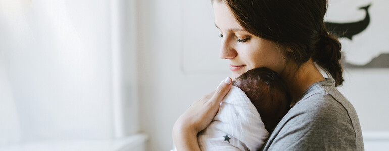 Eine Frau, die ein in ein Tuch gewickeltes Baby im Arm hält, steht in einem hellen Raum an einem Fenster.