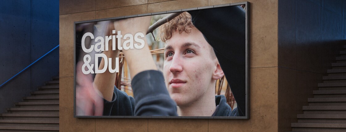 Ein Billboard in einer urbanen Passage zeigt ein Plakat der Caritas, auf dem das Caritas Logo sowie das Portrait eines Mannes zu sehen ist.