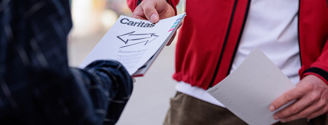 Unternehmen und Kooperationen Ein Caritas Mitarbeiter, der eine rote Jacke trägt, teilt einer Passantin eine Publikation aus.