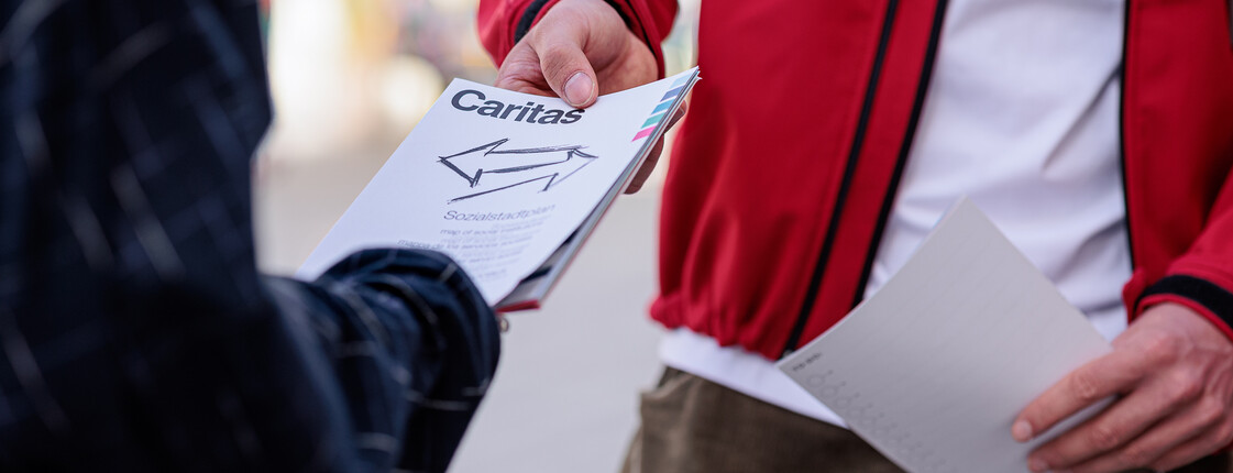 Ein Caritas Mitarbeiter, der eine rote Jacke trägt, teilt einer Passantin eine Publikation aus.
