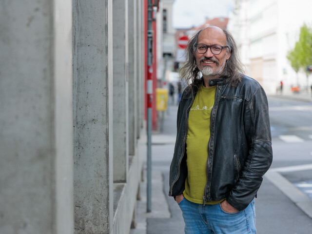 Portrait eines Caritas Mitarbeiters, der neben einer Gebäudeaußenfassade aus Beton steht und die Hände in den Hosentaschen hat.