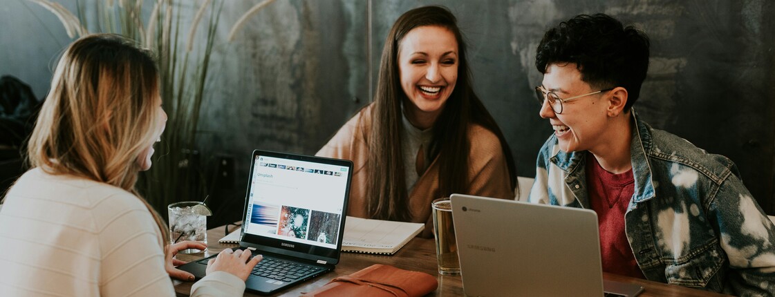 Lernhilfe in den Fächern Mathematik und Englisch Drei Frauen sitzen an einem Holztisch vor ihren Laptops und lachen zusammen.
