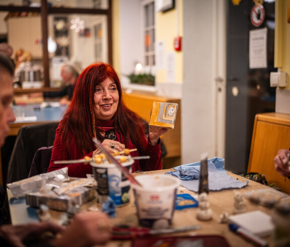 Eine Frau, die mit anderen Personen an einem Tisch sitzt, zeigt ein selbstgemaltes Bild in die Runde.