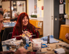 Eine Frau, die mit anderen Personen an einem Tisch sitzt, zeigt ein selbstgemaltes Bild in die Runde.