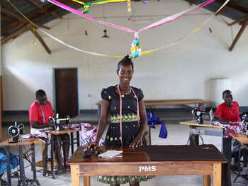Portrait einer jungen Frau aus Uganda, die in einer Halle vor einem Holztisch steht.