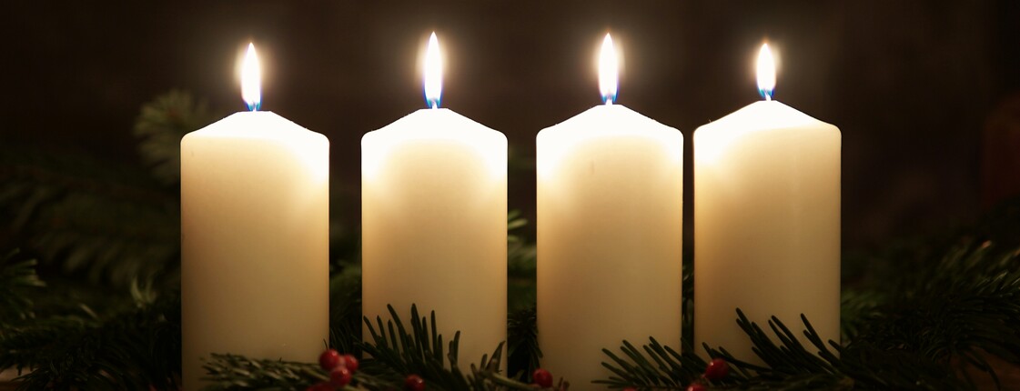 Gute Geschichten im Advent Vier weiße, brennende Kerzen stehen nebeneinander auf einem Adventkranz.