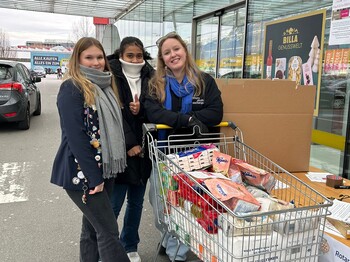 Drei Frauen in Winterkleidung präsentieren vor einem Supermarkt einen vollen, als Spende vorgesehenen Einkaufswagen.