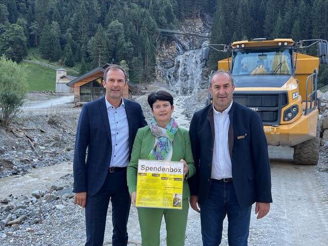 Drei Personen, zwei Männer in Anzügen und eine Frau in grünem Anzug, stehen vor dem vom Unwetter zerstörten Mühlendorf in Gschnitz.
