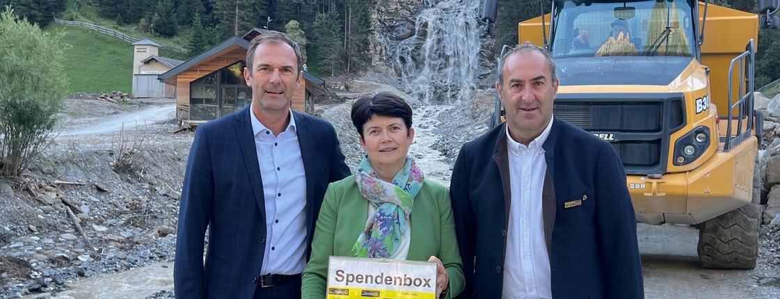 Katastrophenhilfe Gschnitz  Drei Personen, zwei Männer in Anzügen und eine Frau in grünem Anzug, stehen vor dem vom Unwetter zerstörten Mühlendorf in Gschnitz.