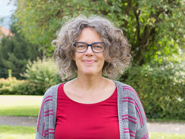 Portrait einer Frau mit rotem T-Shirt und Brille, die in einem Garten steht.