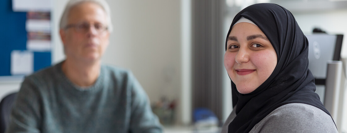 Flucht, Migration und Integration Portrait einer jungen Frau mit schwarzem Kopftuch während eines Beratungsgesprächs. Im Hintergrund sitzt ein Caritas Mitarbeiter an einem Schreibtisch.