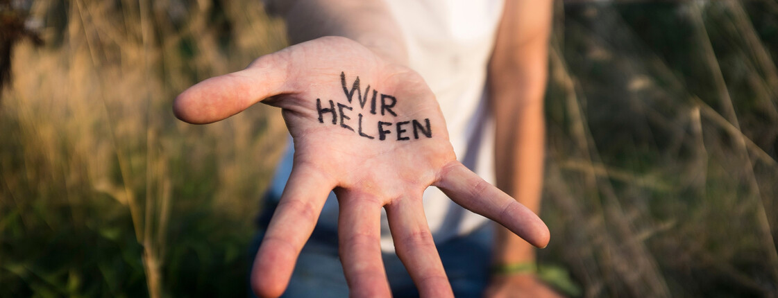 Eine Person, die ein weißes T-Shirt trägt und in einer Sommerwiese steht, streckt eine Hand mit Schriftzug "Wir helfen" in die Kamera.