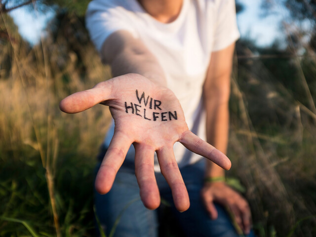 Eine Person, die ein weißes T-Shirt trägt und in einer Sommerwiese steht, streckt eine Hand mit Schriftzug "Wir helfen" in die Kamera.