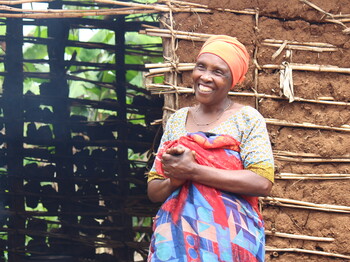 Portrait einer lachenden Frau in traditioneller Kleidung in Tansania, die vor einer Lehmhütte steht.