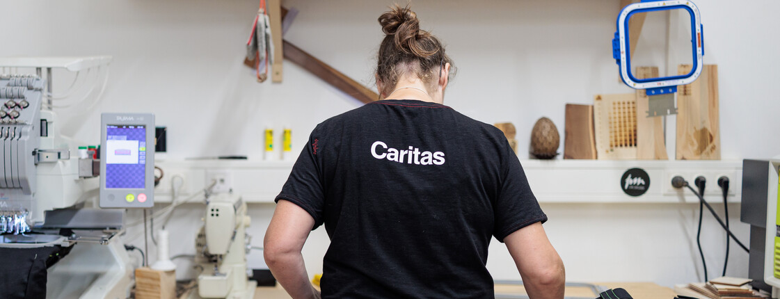 Eine Frau, die ein schwarzes T-Shirt mit weißem Caritas-Logo trägt, steht mit dem Rücken zur Kamera an einer Werkbank aus Holz. 