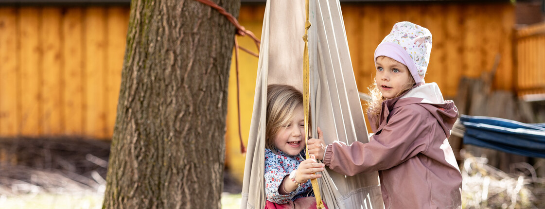 Zwei Kindergartenkinder in Regenkleidung spielen im Garten mit einer Stoff-Schaukel.
