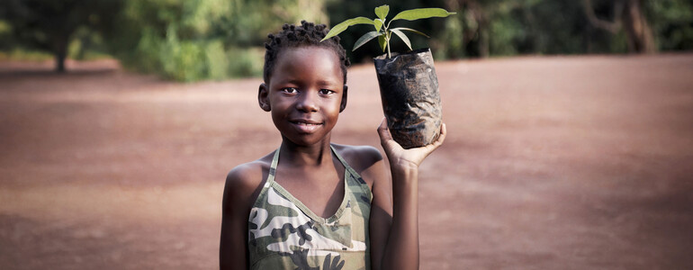 Portrait eines Kindes in Afrika, das einen Pflanzensetzling in der linken Hand hoch hält.