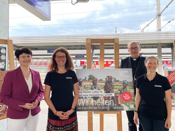 Vier Personen stehen auf einem Bahnsteig und präsentieren Plakate zur Caritas Spendenaktion "Gegen den Hunger"