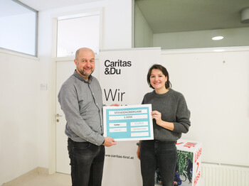Ein Mann und eine Frau mittleren Alters stehen vor einem Caritas Roll-Up und präsentieren einen Spendenscheck in Höhe von 2.000 €.
