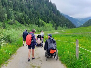 Eine Gruppe, darunter Menschen mit Behinderung, wandert einen Almweg in einem Bergtal entlang.