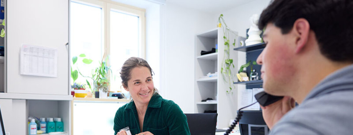Mitarbeiterin spricht im JUCA-Büro mit jungem Erwachsenen, der telefoniert; Gesprächssituation mit Computer am Schreibtisch.