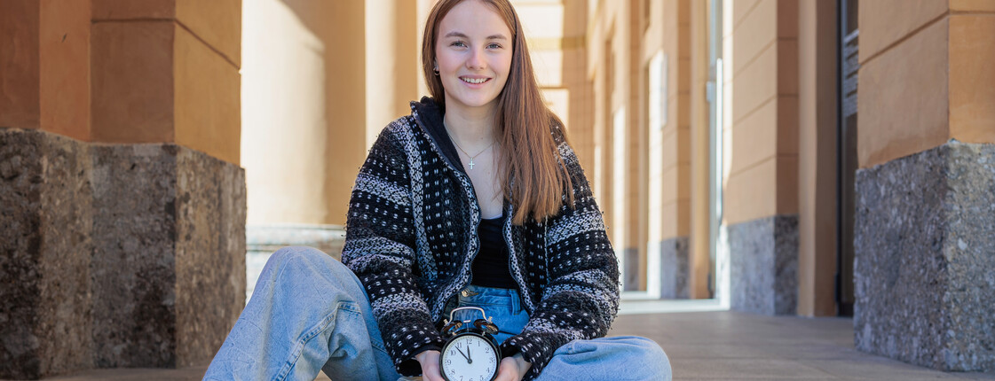 Portrait einer Schülerin in Streetwear, die im Schneidersitz auf einer Betontreppe sitzt und einen großen Wecker in den Händen hält.