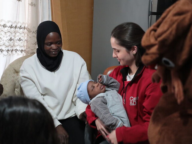 Eine Frau mit roter Caritas-Jacke hält ein Baby in Händen. Die Mutter des Babys sitzt daneben.