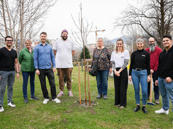 Neun Personen stehen auf einer grünen Wiese um einen frisch gepflanzen Baum zu einem Gruppenfoto zusammen.