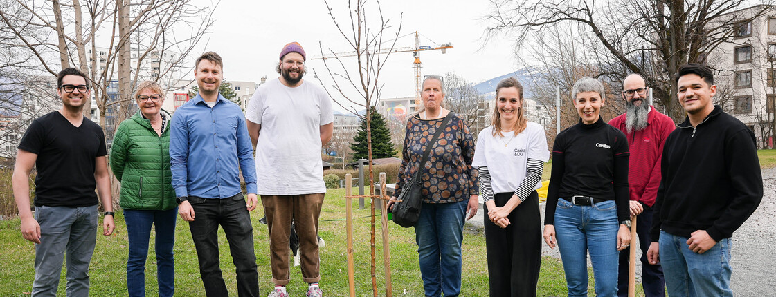 Neun Personen stehen auf einer grünen Wiese um einen frisch gepflanzen Baum zu einem Gruppenfoto zusammen.