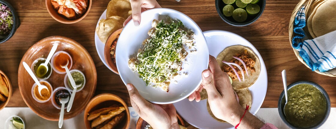 Vogelperspektive eines voll gedeckten Essenstisch aus Holz, an dem Personen sitzen und sich gegenseitig Schüsseln mit Essen reichen.