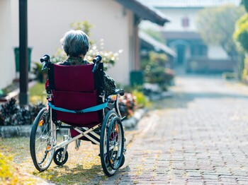 Workshop: Achtsame Begleitung von Menschen mit Demenz Eine im Rollstuhl sitzende Seniorin befindet sich in einer Hauseinfahrt und schaut die Straße hinunter.