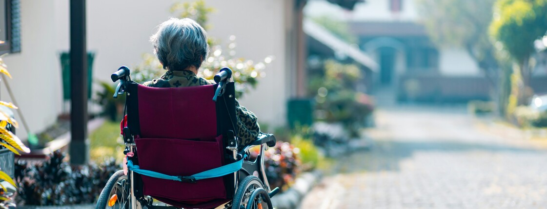 Eine im Rollstuhl sitzende Seniorin befindet sich in einer Hauseinfahrt und schaut die Straße hinunter.