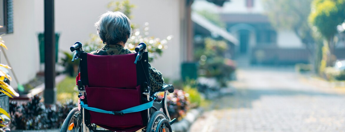 Eine im Rollstuhl sitzende Seniorin befindet sich in einer Hauseinfahrt und schaut die Straße hinunter.