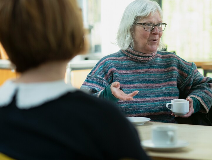 Bei einem Angehörigencafé erzählt eine Seniorin mit Brille, die eine Kaffeetasse in der Hand hält, den anderen Teilnehmerinnen von ihrem Alltag. 