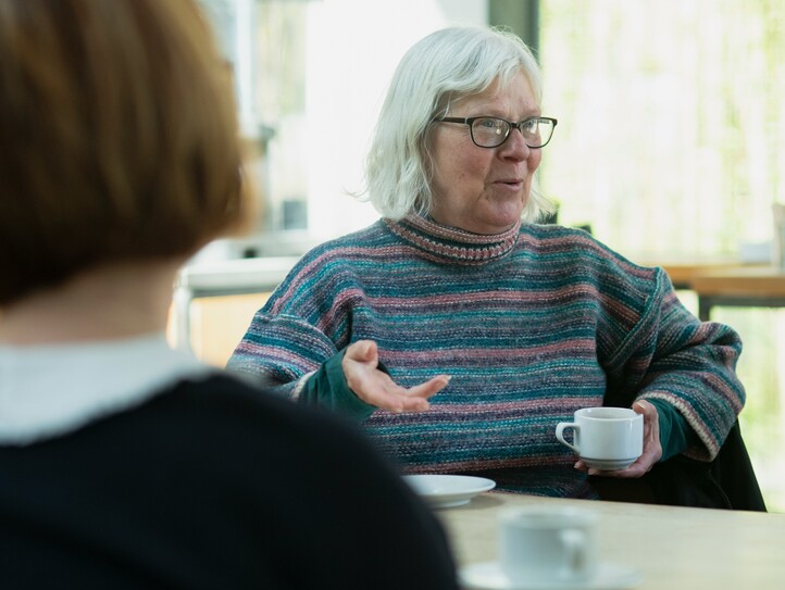 Bei einem Angehörigencafé erzählt eine Seniorin mit Brille, die eine Kaffeetasse in der Hand hält, den anderen Teilnehmerinnen von ihrem Alltag. 