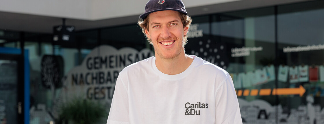Einer junger Mann mit Shildkappe und weißem Caritas T-Shirt sitzt auf einer Bank vor einem Gebäude.