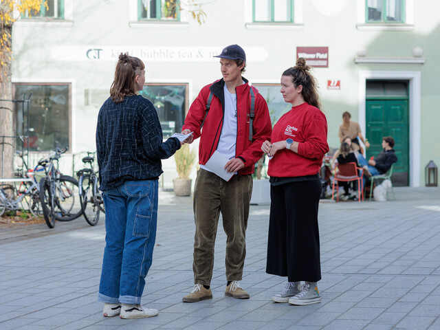 Zwei Caritas Mitarbeiter*innen stehen auf einem kleinen Platz und verteilen einen Informationsbroschüre an eine Passantin.