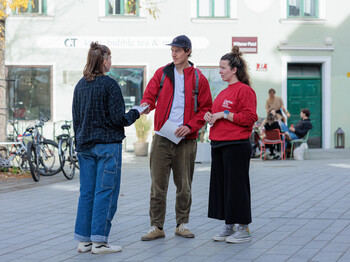 Zwei Caritas Mitarbeiter*innen stehen auf einem kleinen Platz und verteilen einen Informationsbroschüre an eine Passantin.