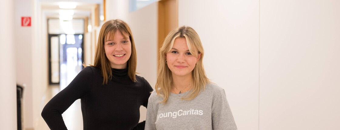 Portrait zweier junger Frauen, die nebeneinander in einem hell erleuchteten Gang stehen.