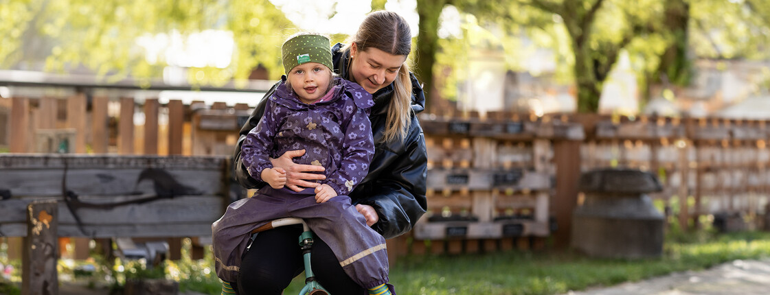 Eine Frau mittleren Alters und ein Mädchen in Regenkleidung fahren in einem Garten mit einem Kinderfahrrad.