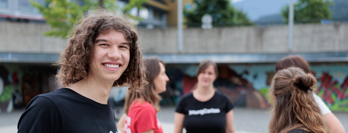 Portrait eines jungen Mannes mit schwarzem T-Shirt. Im Hintergrund unterhalten sich vier Personen im Kreis stehend.