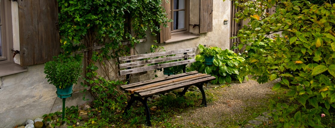 Eine alte Holzbank steht im Garten an der Hauswand eines alten, malerischen Hauses.