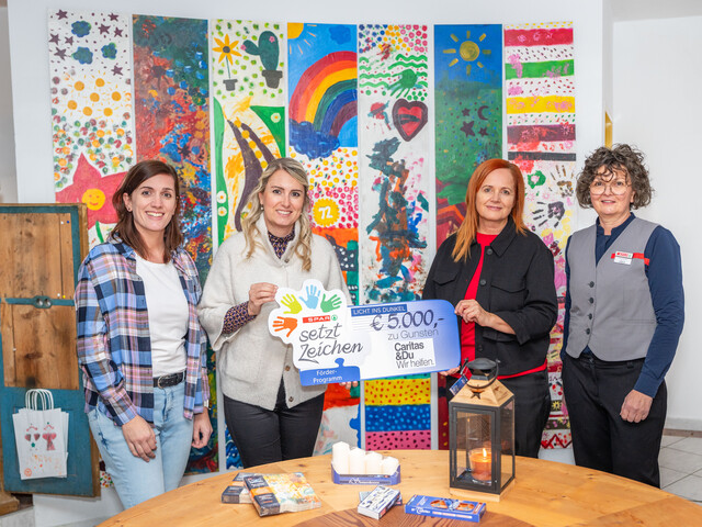 Vier Frauen stehen vor einer bunt bemalten Fotowand und präsentieren einen Spendenscheck in Höhe von 5.000 Euro.
