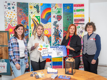 Vier Frauen stehen vor einer bunt bemalten Fotowand und präsentieren einen Spendenscheck in Höhe von 5.000 Euro.