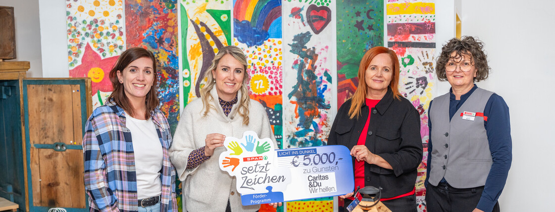 Vier Frauen stehen vor einer bunt bemalten Fotowand und präsentieren einen Spendenscheck in Höhe von 5.000 Euro.