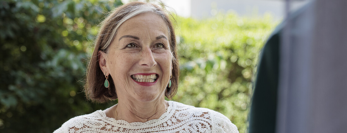 Eine Haussammlerin, die eine Filztragetasche und ein rotes Klemmbrett in den Händen hält, steht lächelnd an einer geöffneten Eingangstür.