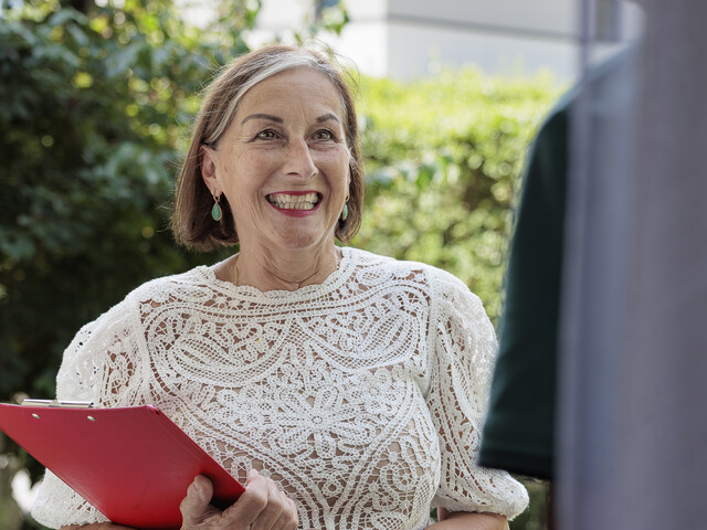 Eine Haussammlerin, die eine Filztragetasche und ein rotes Klemmbrett in den Händen hält, steht lächelnd an einer geöffneten Eingangstür.
